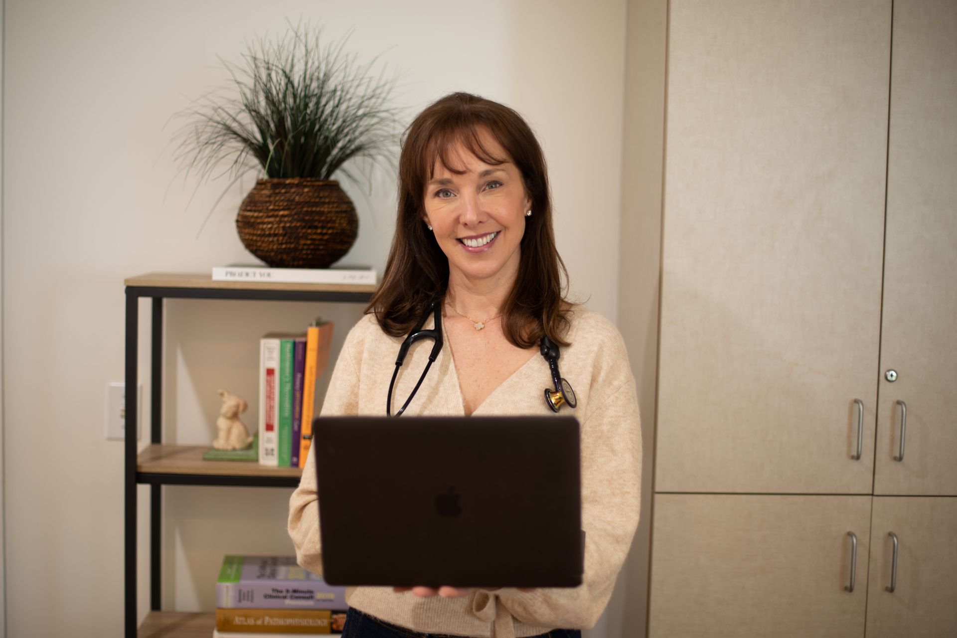 Woman with a stethoscope holds a laptop, smiling at the camera in an office setting.