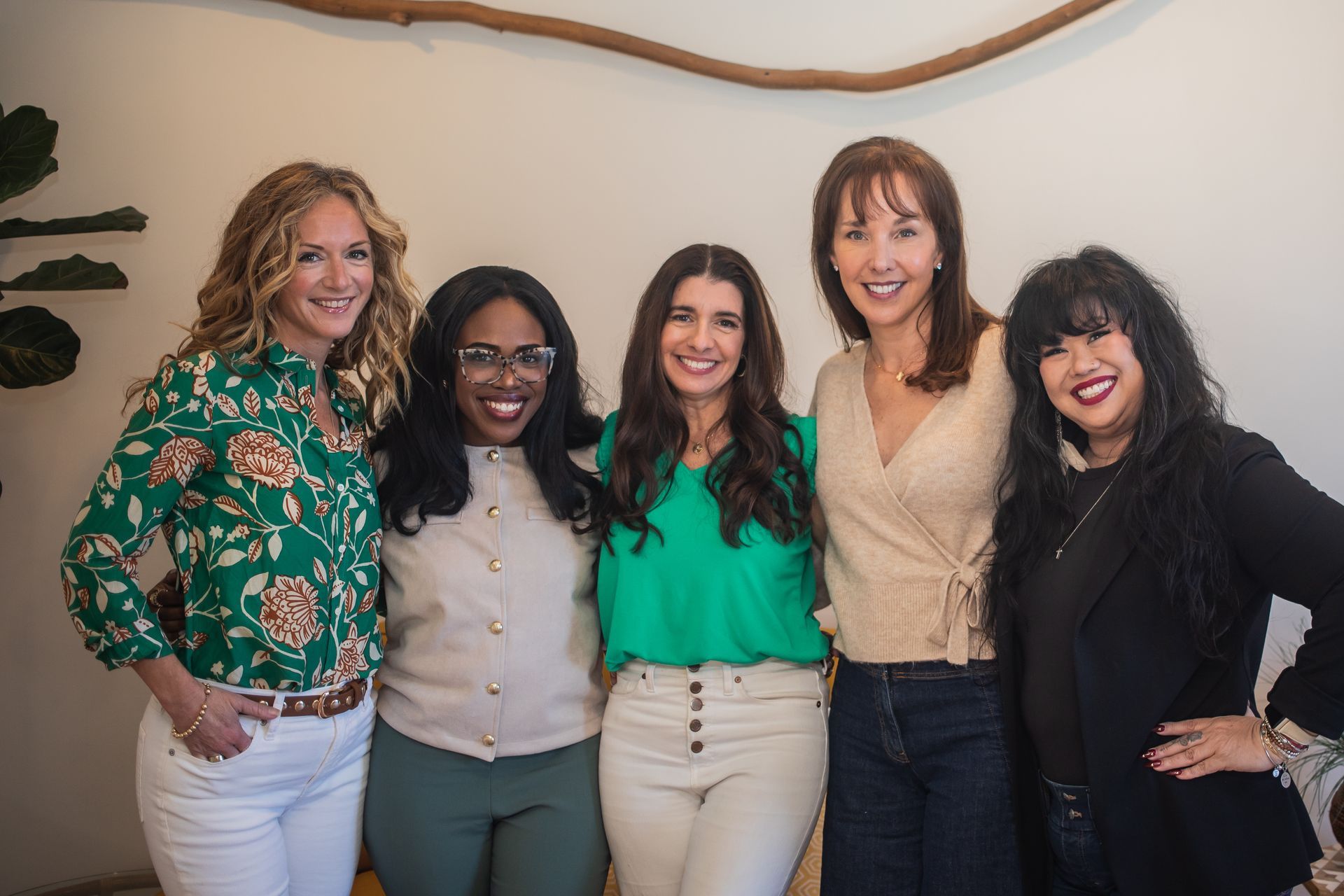 Five women smiling, posing closely together against a white wall with a decorative branch and plant.
