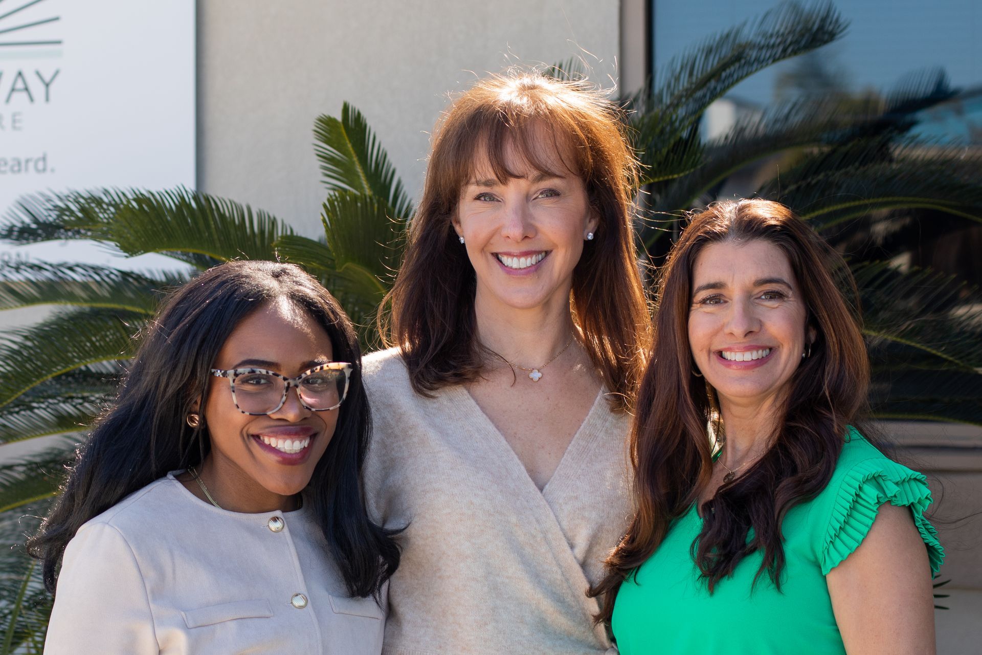 Three smiling women pose outdoors in front of a building and foliage; one wears glasses, another a green top.