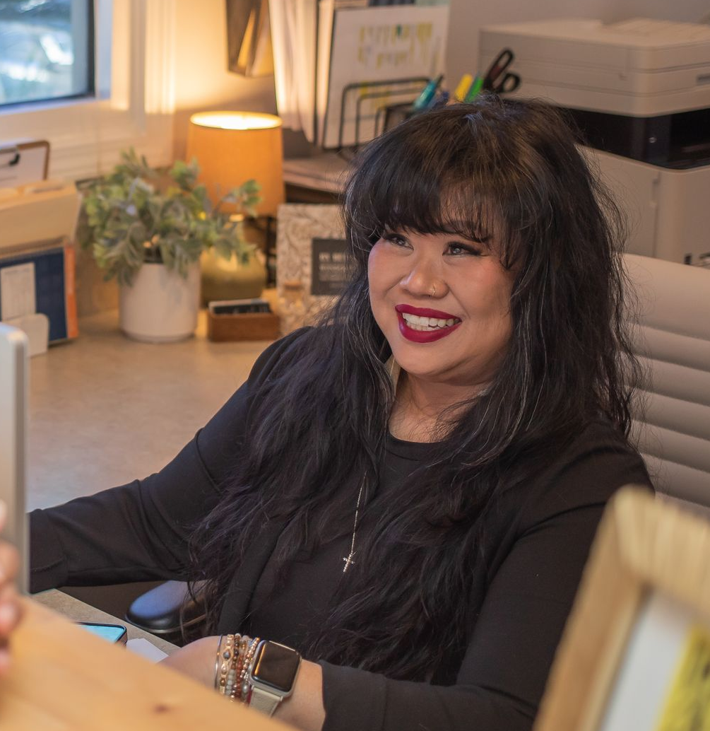 Woman with dark hair smiles while seated at a desk. Office setting, lamp, and other office supplies are visible.