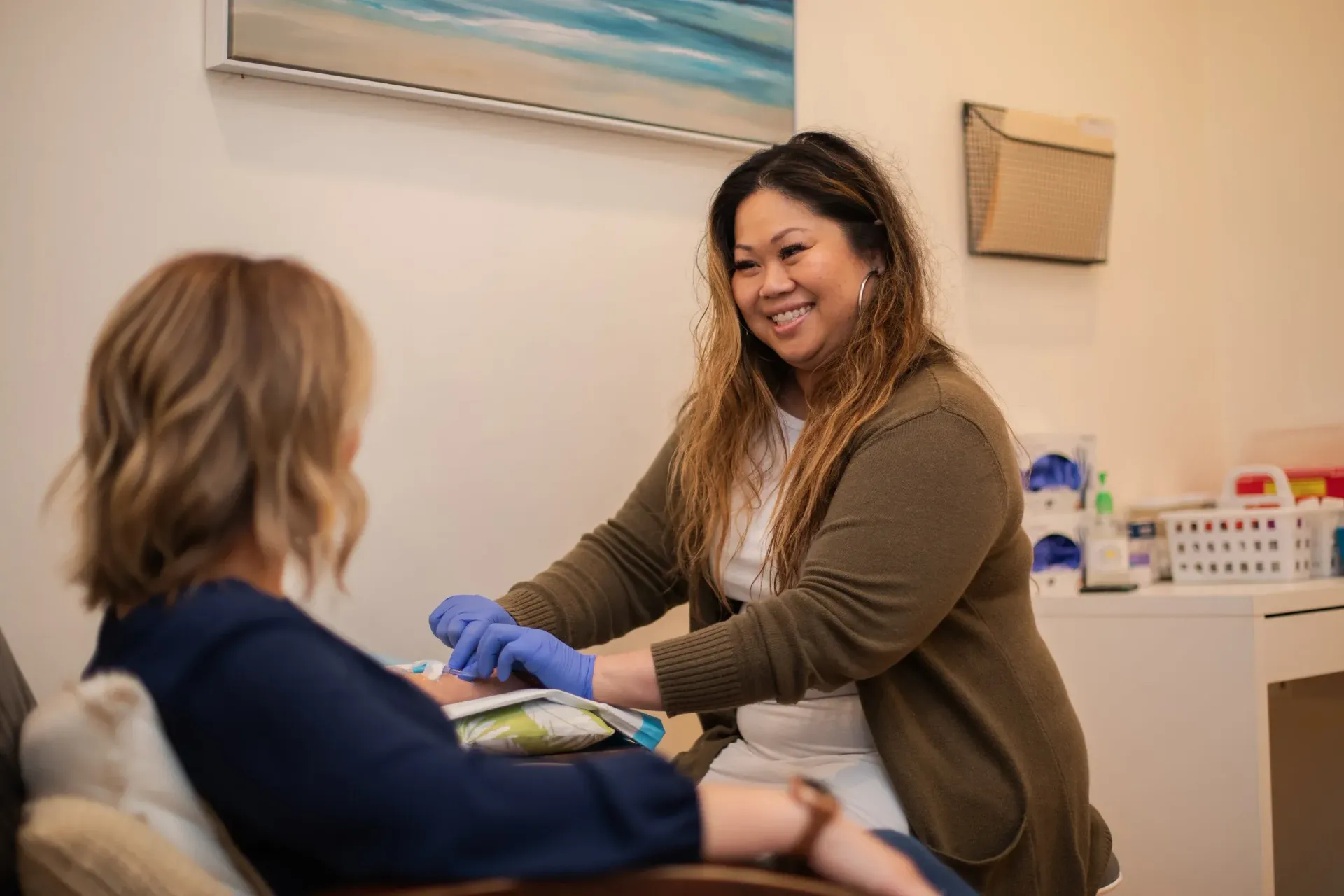 A woman in a clinic prepares to draw blood from a patient, both smiling. The setting is bright and clean.