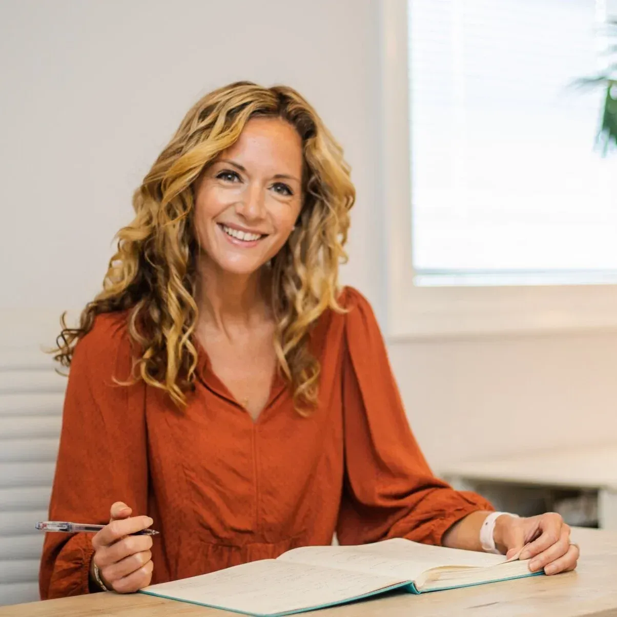 Woman with curly blonde hair smiles, holding pen, looking at open book on a desk. Room with window.