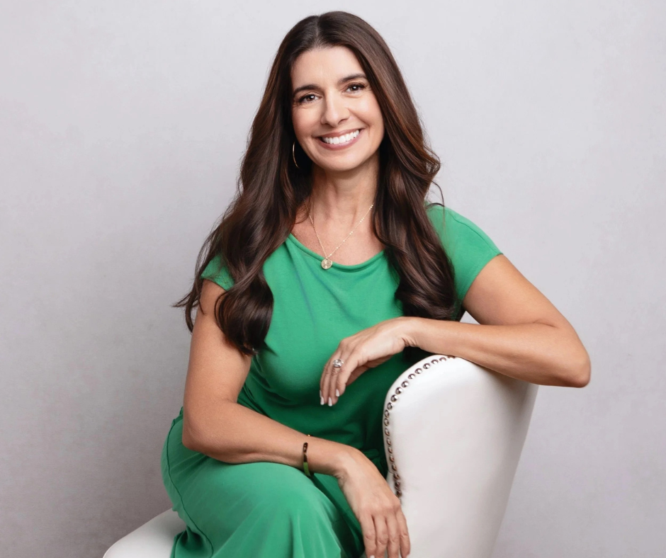 Woman with long brown hair, smiling, wearing a green dress, sitting on a white chair against a neutral backdrop.