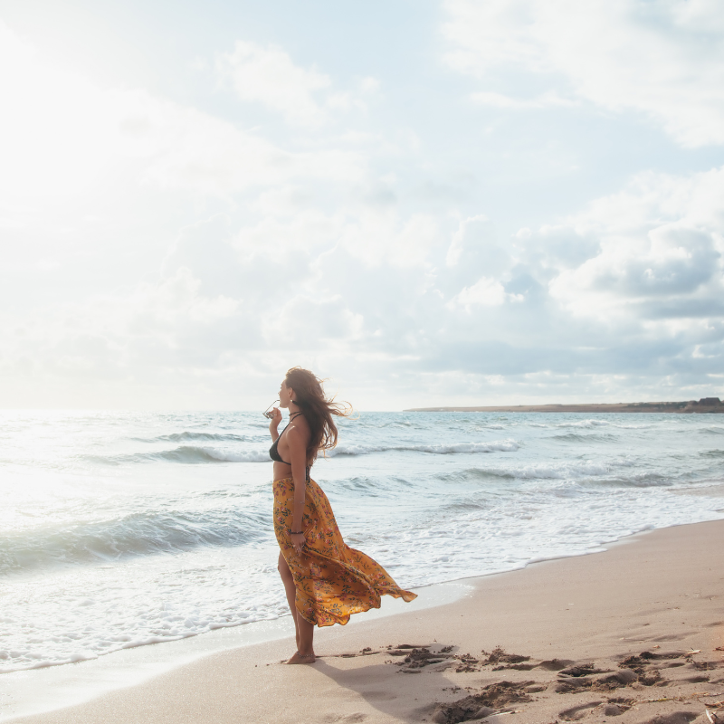 A person in a flowing patterned skirt stands on a sandy beach, gazing out at the ocean under a cloudy sky.