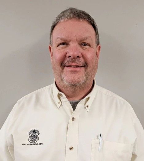 A man with a beard is wearing a white shirt and smiling.