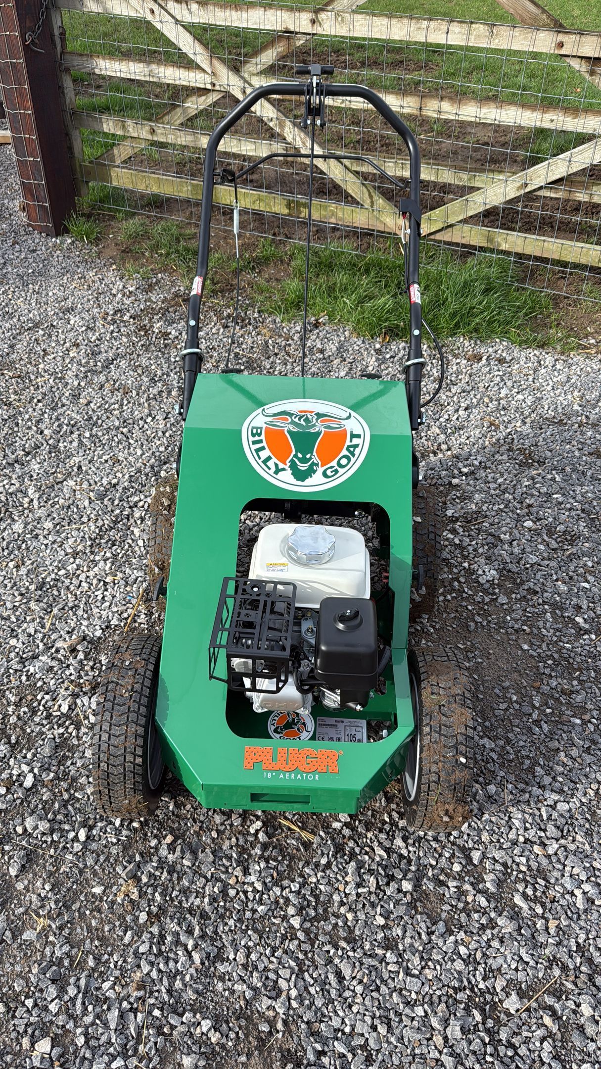 Green and white lawn scarifier on gravel, with a brick wall and wooden fence in background.