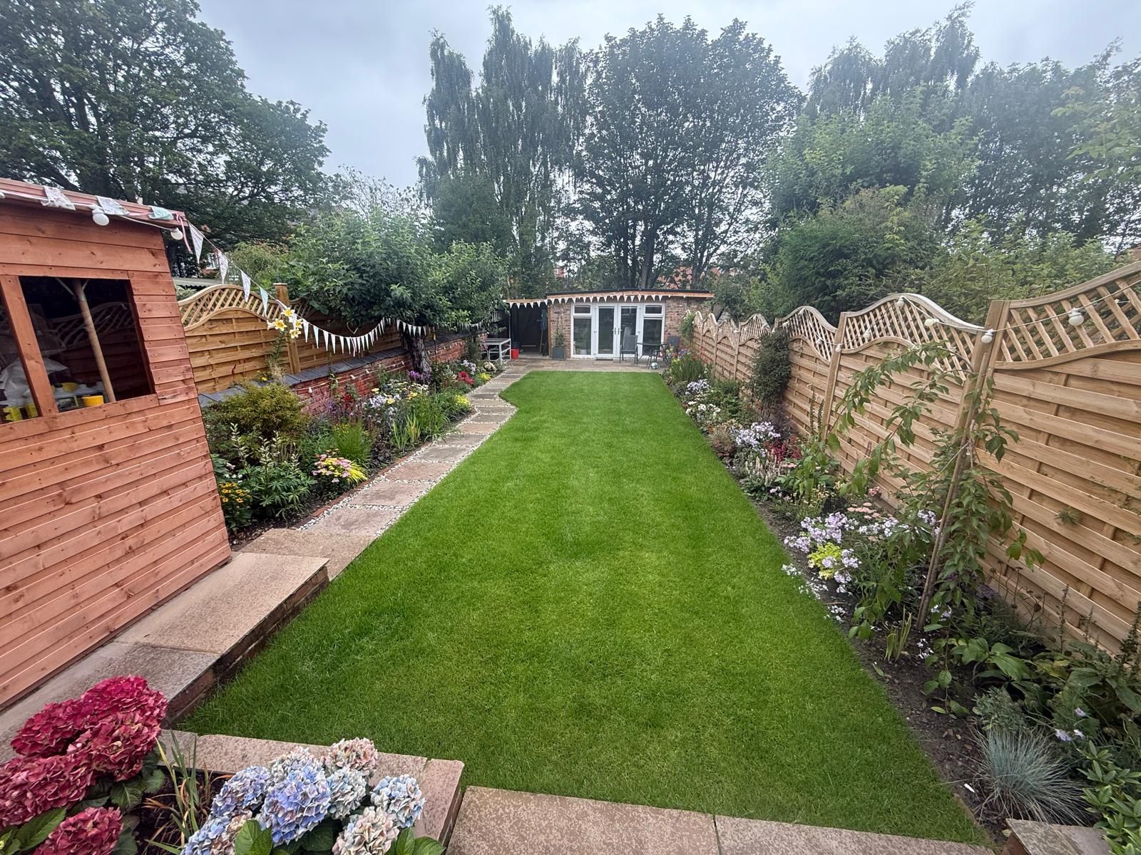 Lush green backyard with a grassy path, flower beds, wooden fences, and a shed.