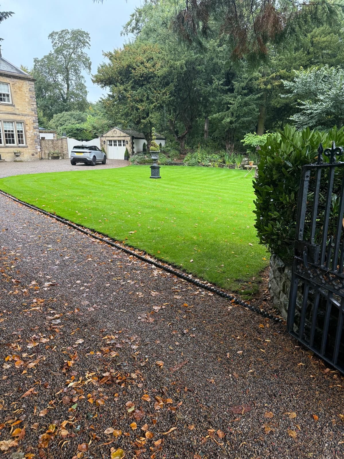 A well-manicured lawn with a gravel driveway leading to a stone house and a garage, surrounded by trees.