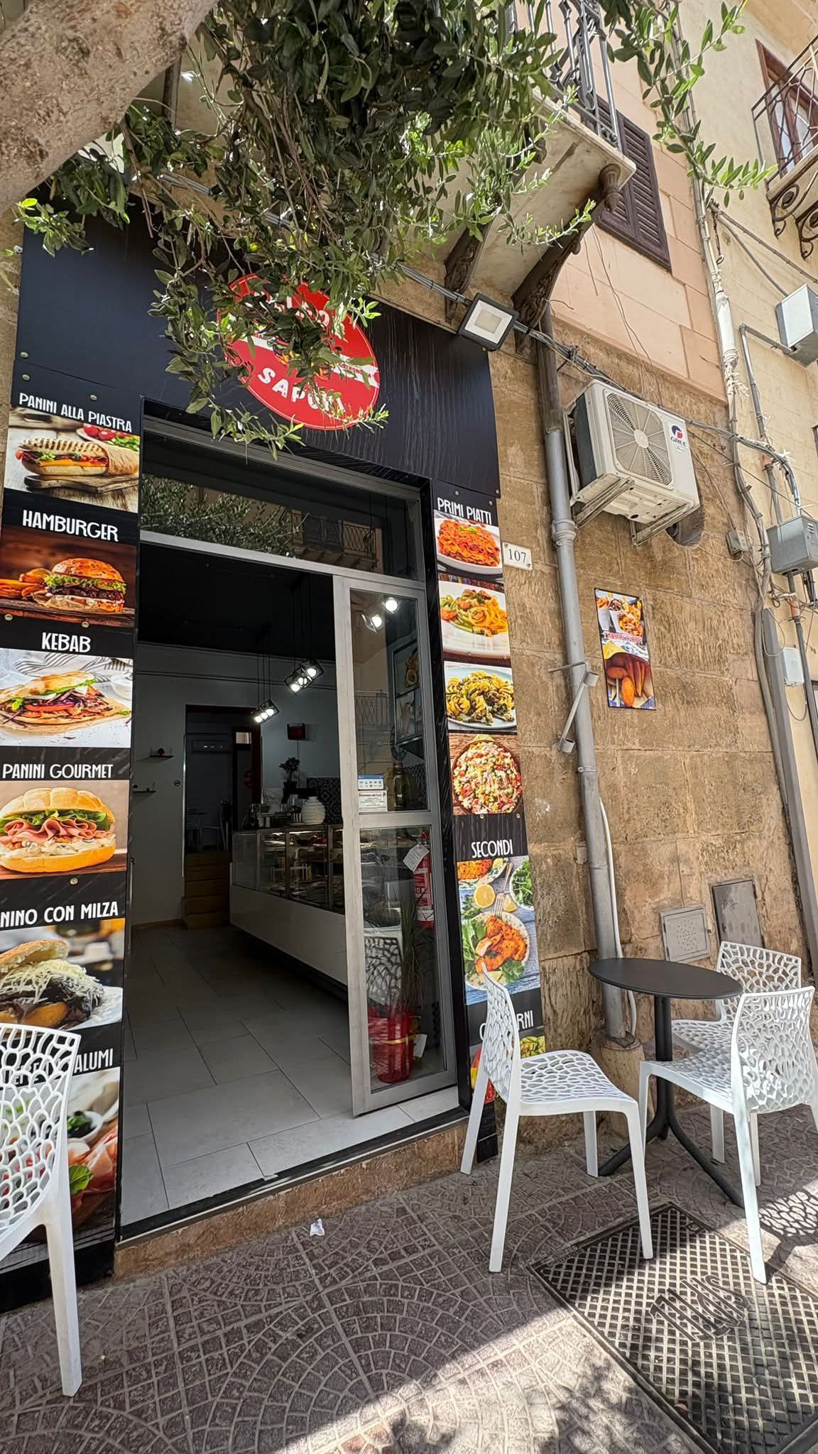 Piccolo ingresso di un caffè con porta aperta, cartelli con il menù, piante sospese e tavolini all'aperto su una strada lastricata.