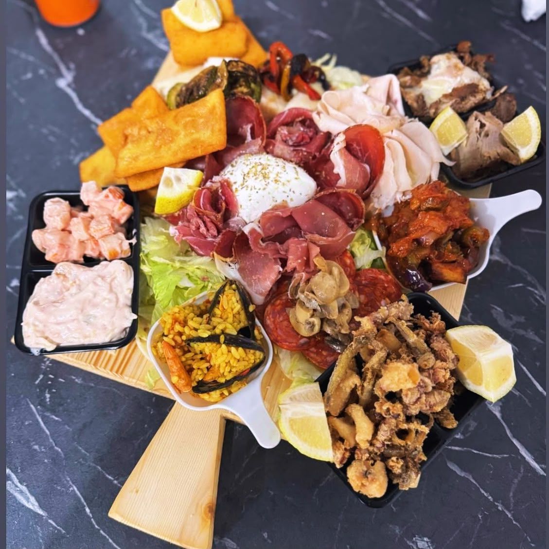 Piatto misto di frutti di mare e tapas con spicchi di limone, salumi e fritture su un tavolo scuro.