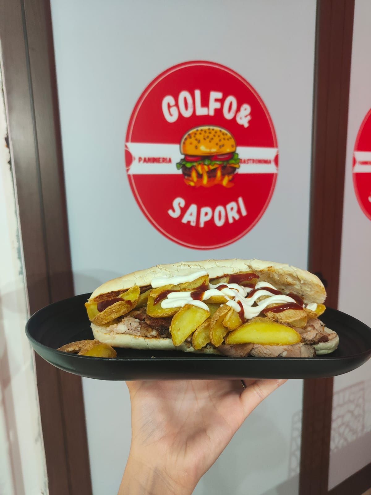 Una mano che regge un piatto nero con un panino e patatine fritte davanti a un'insegna rossa del Golfo & Sapori.