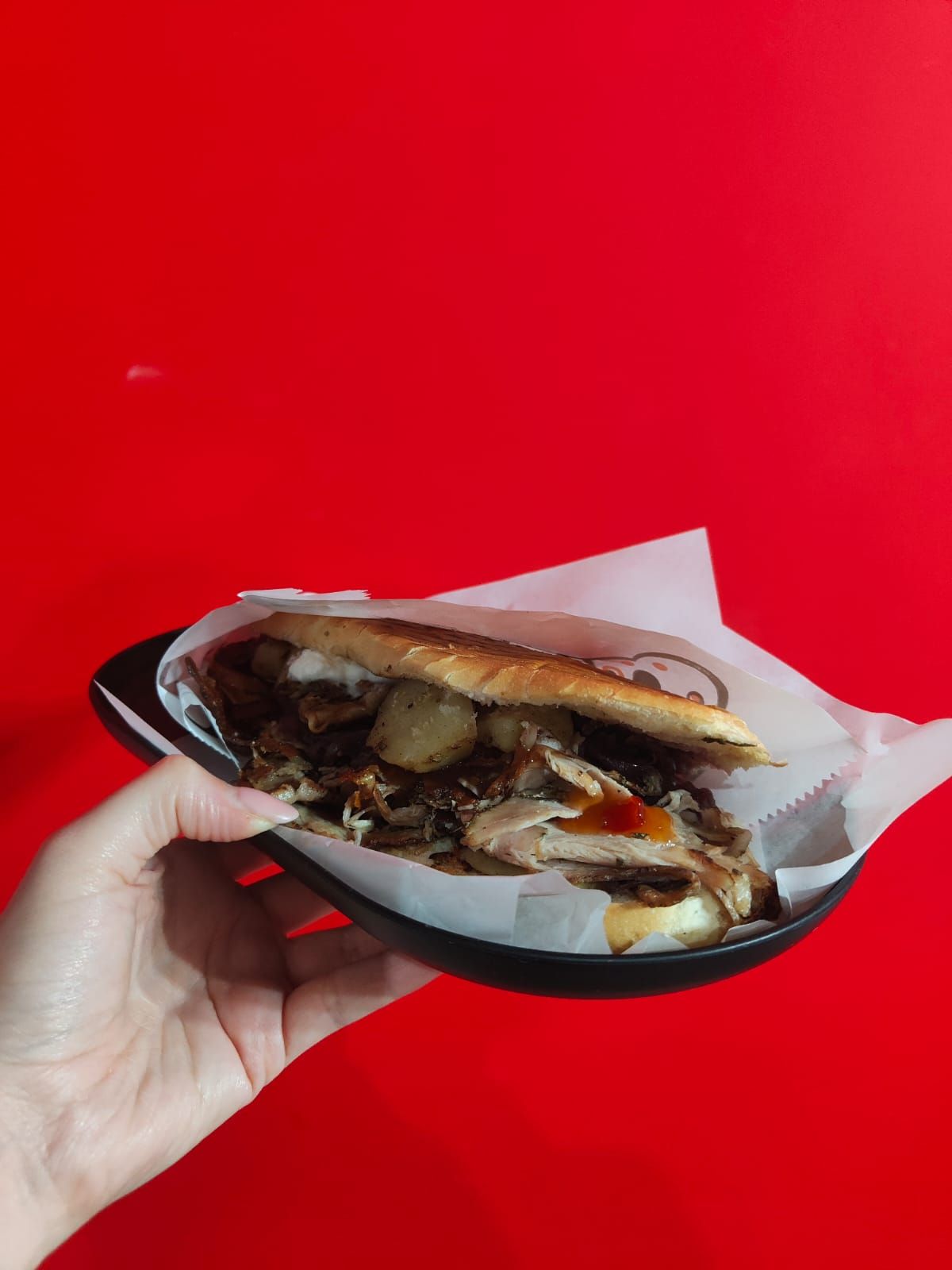 Una mano che regge un piatto nero con un panino, su uno sfondo rosso acceso.