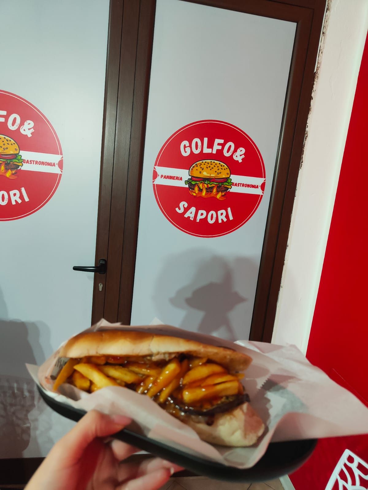 Una mano che regge un panino davanti a una porta bianca con il logo rosso
