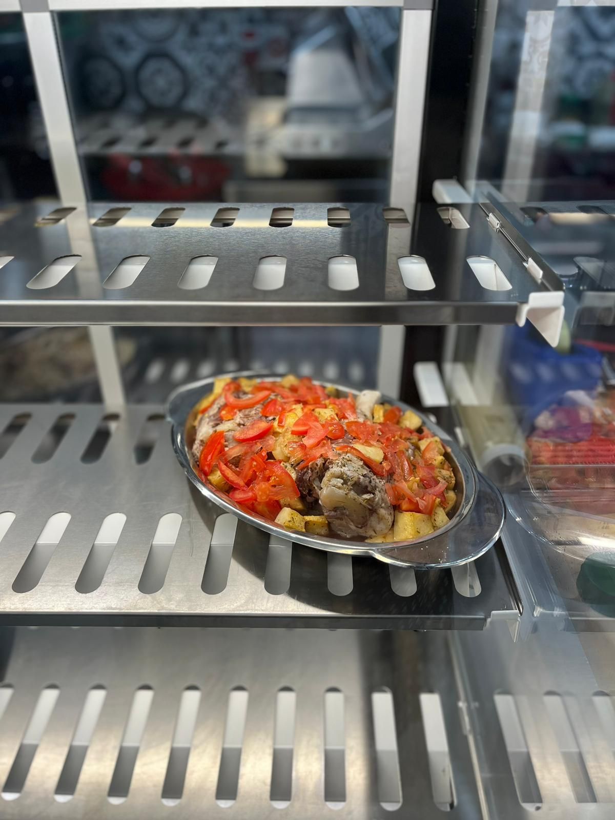 Un vassoio di alluminio contenente cibo al forno guarnito con peperoni rossi è appoggiato su uno scaffale metallico all'interno di una vetrina.