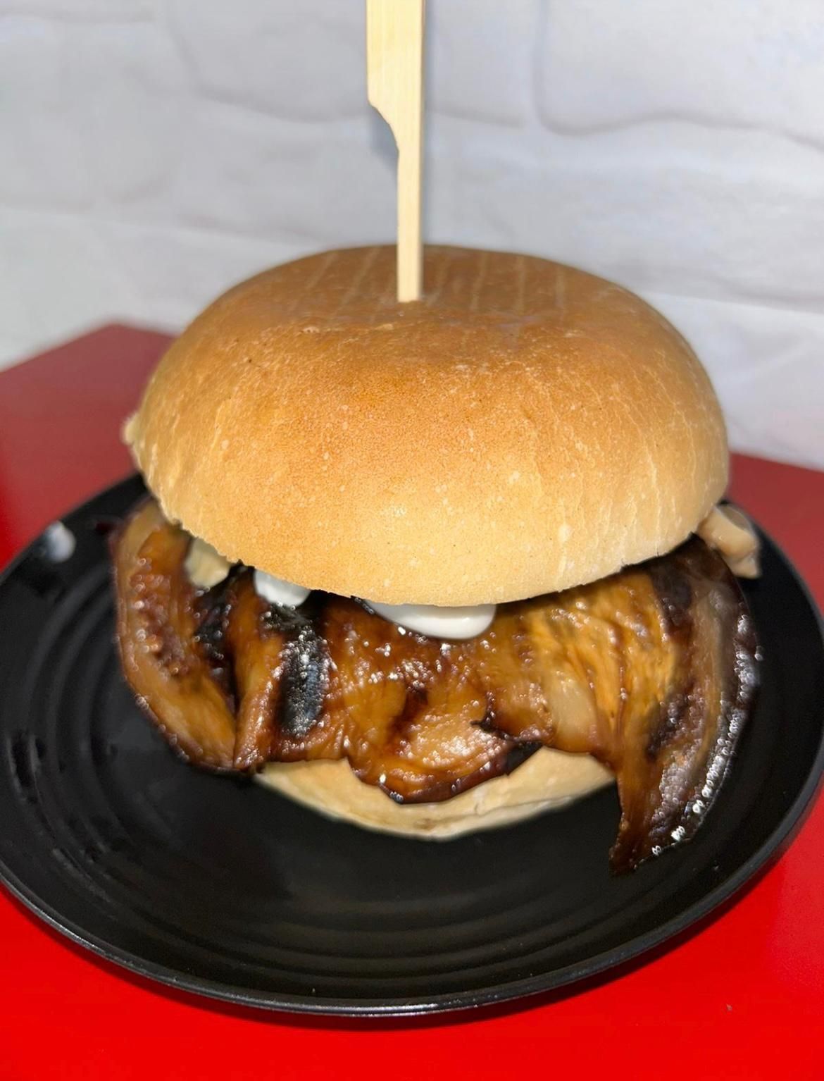 Panino con pollo grigliato su un piatto nero con vassoio rosso, sormontato da un panino e uno spiedino di legno.
