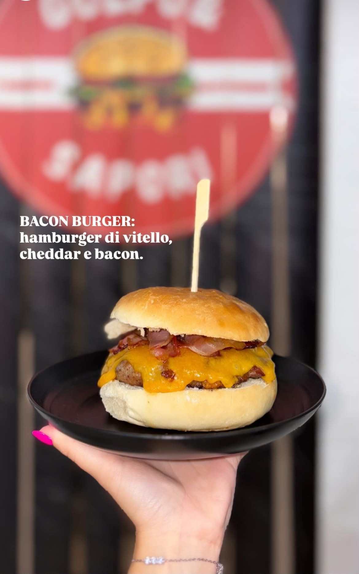 Un cheeseburger con pancetta su un piatto nero tenuto in mano, con l'insegna rossa di un ristorante sfocata sullo sfondo.