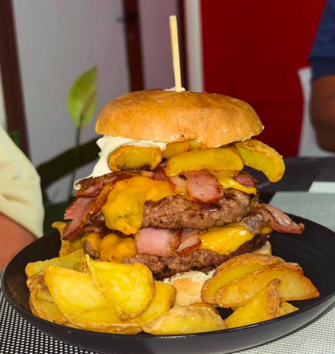 Cheeseburger alto con pancetta e patatine fritte su un piatto nero, sormontato da uno spiedino