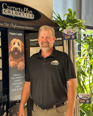 A man in a black shirt is standing in front of a carpets plus color tile display.