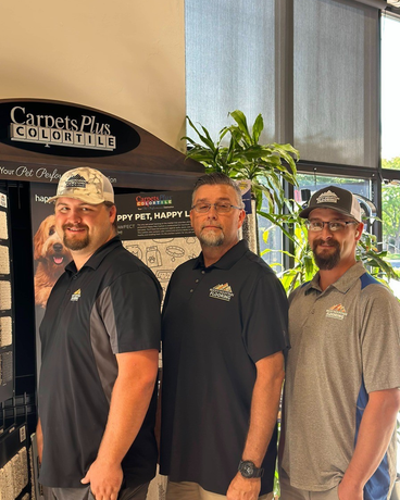 Three men are standing next to each other in front of a sign that says carpets plus schedules.