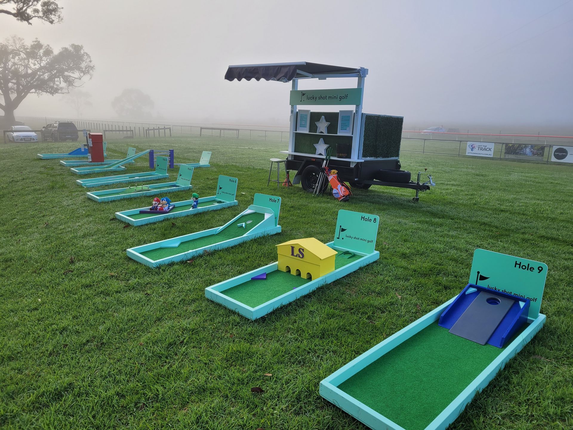 Miniature golf course on a grassy field in foggy conditions, with various obstacles and a food stand.