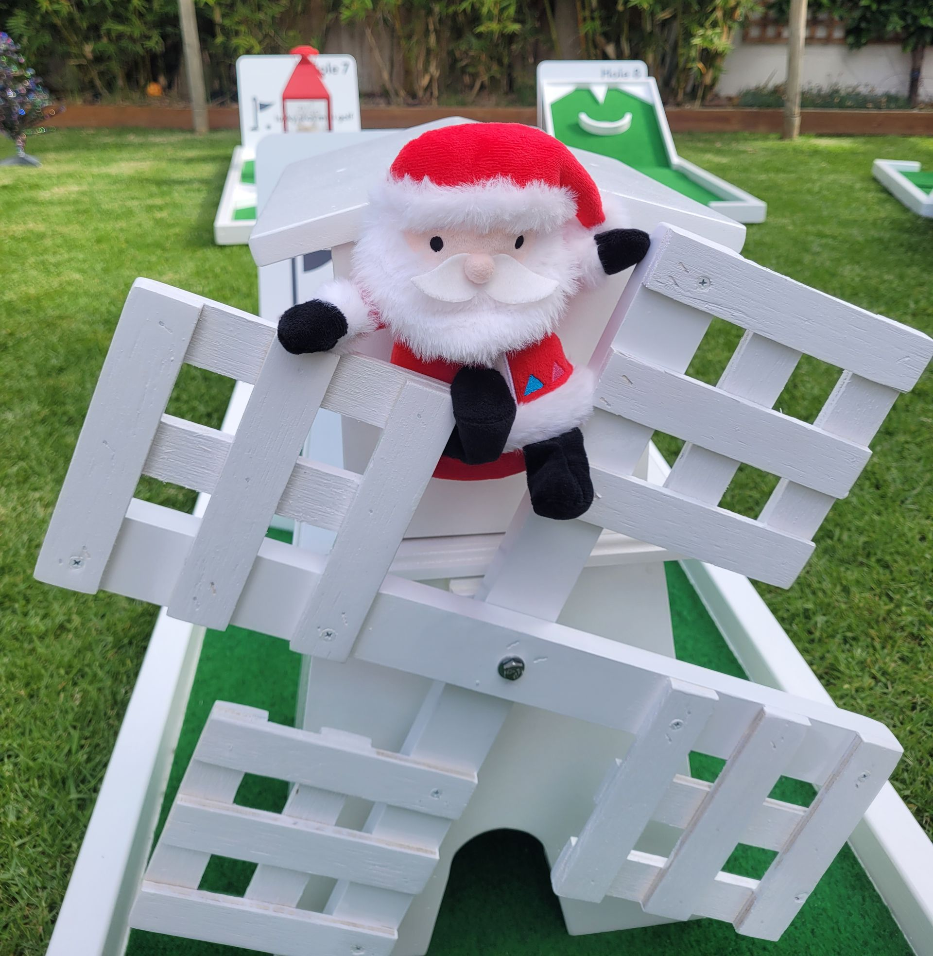 A Santa Claus plush toy sits on top of a white wooden miniature windmill on a mini-golf course.