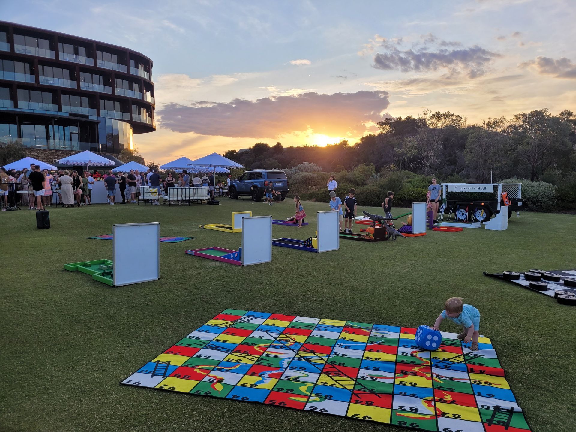 Sunset over an outdoor event with a child playing on a colorful mat. Large building and crowd in background.