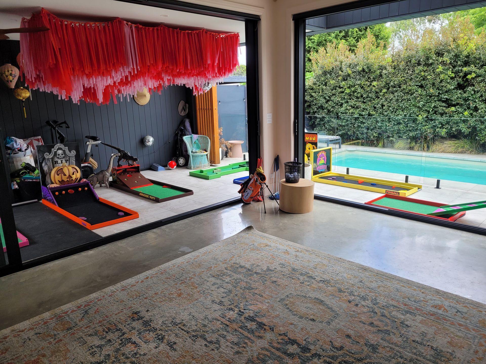 Indoor recreational space with mini golf courses, pool visible through an open doorway, and a rug in the foreground.
