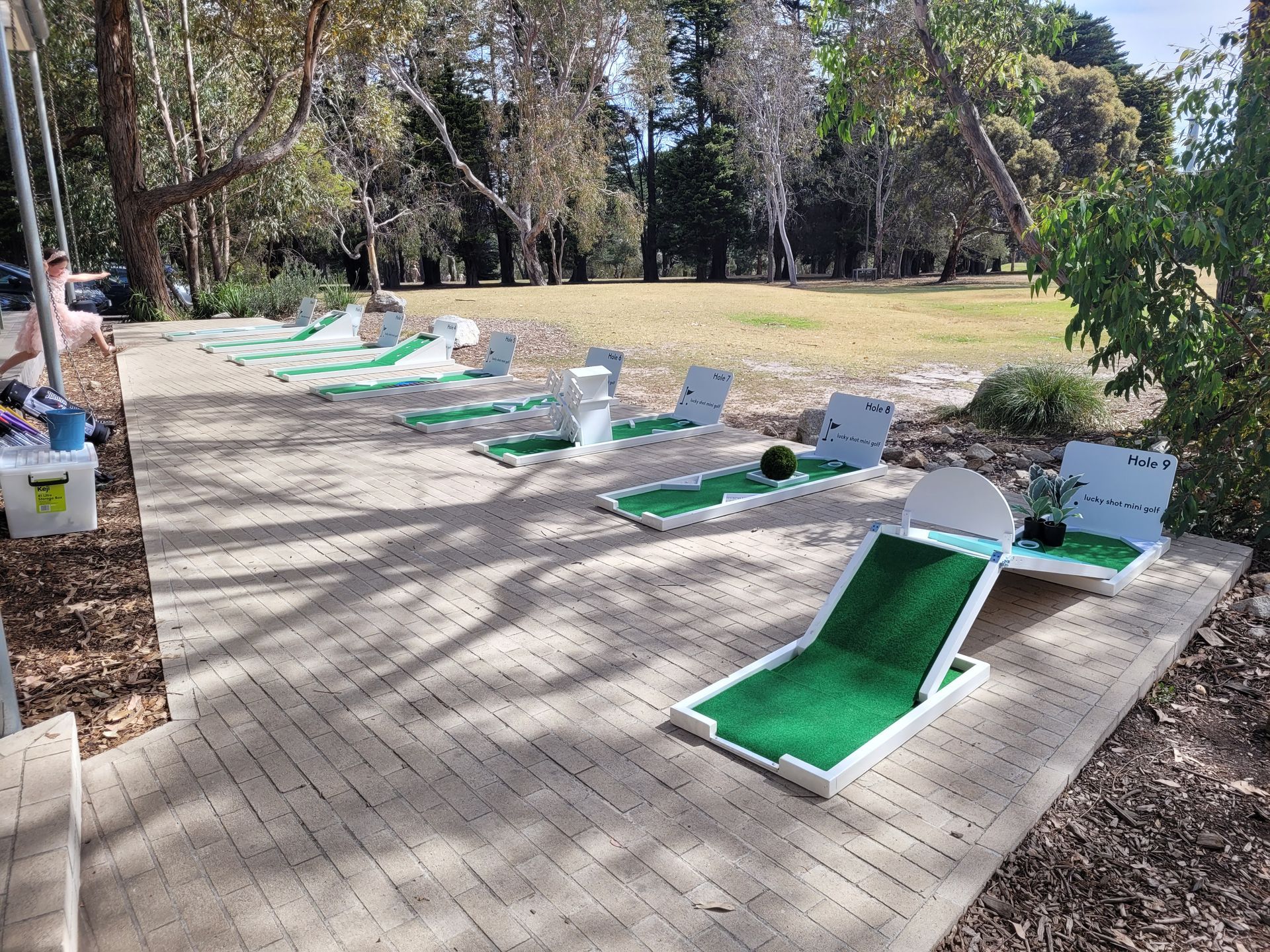 Mini-golf course with green and white structures on brick pavers, set in a park with trees and grass.