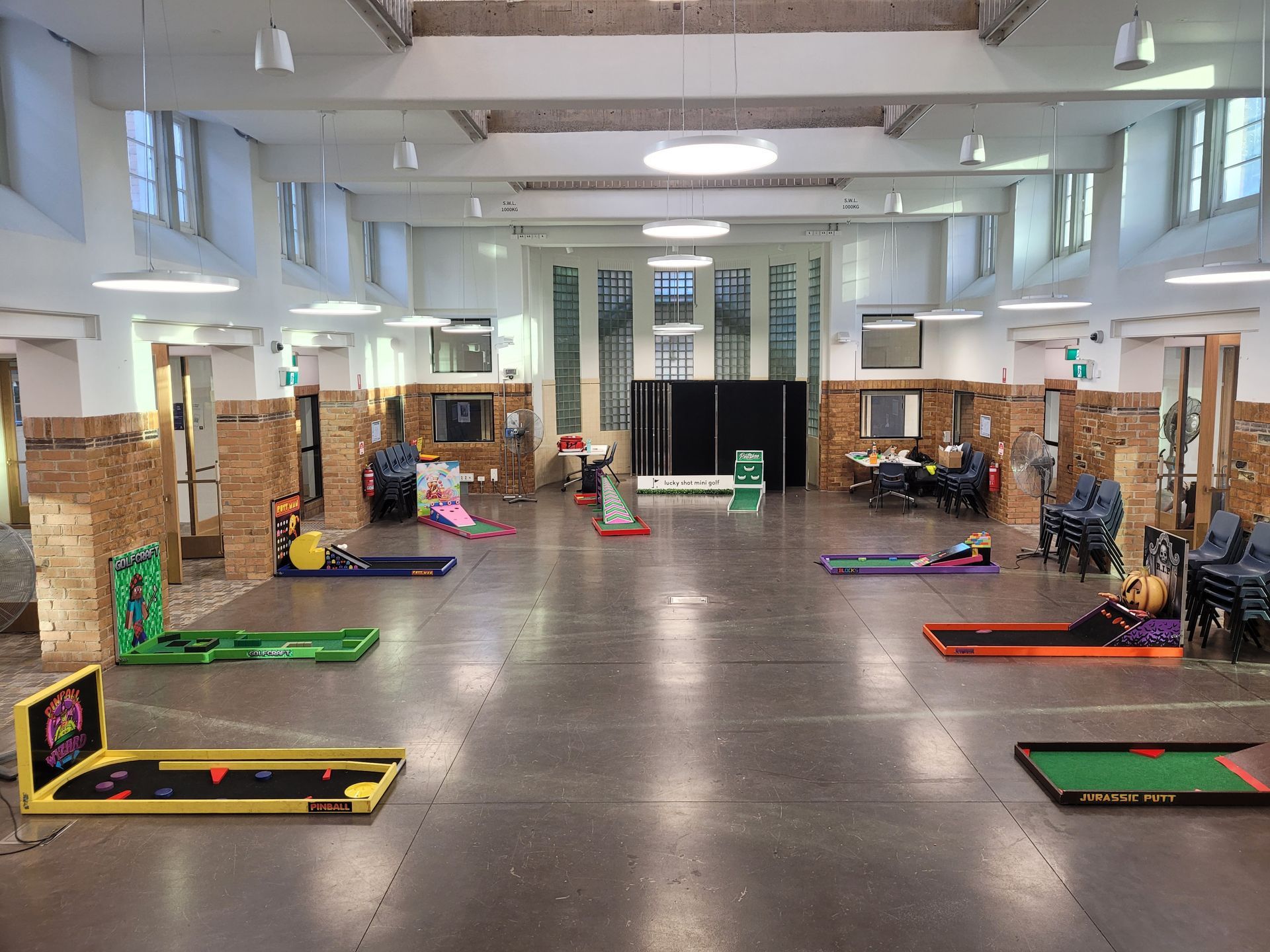 Indoor mini-golf setup with several colorful courses. People sit and play in a spacious hall with brick pillars.