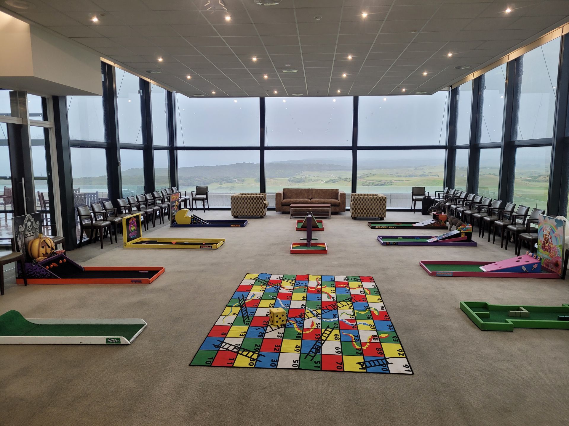 Indoor recreational space with mini golf courses and seating, overlooking a landscape through large windows.