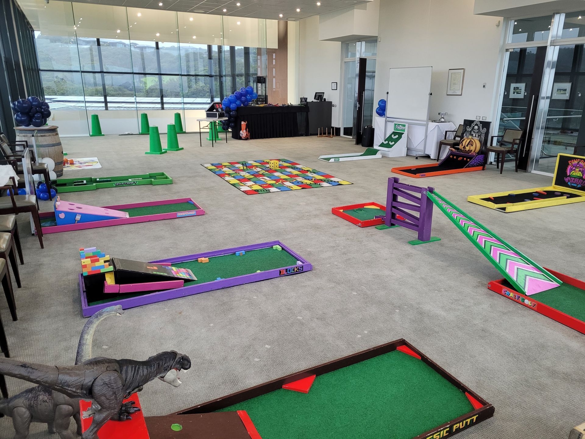 Miniature golf course set up indoors. Several holes with ramps and obstacles, including a dinosaur.