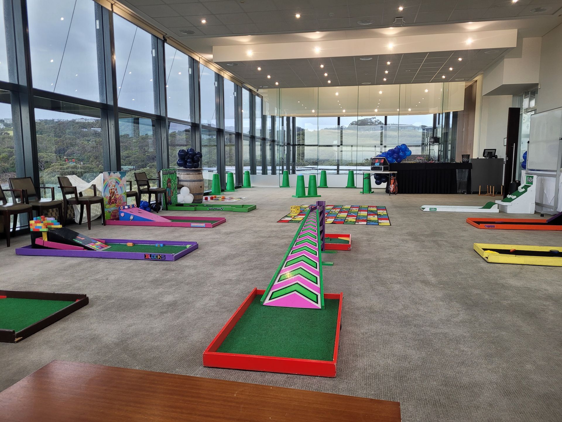 Indoor miniature golf course set up on carpeted floor, with windows overlooking a landscape.