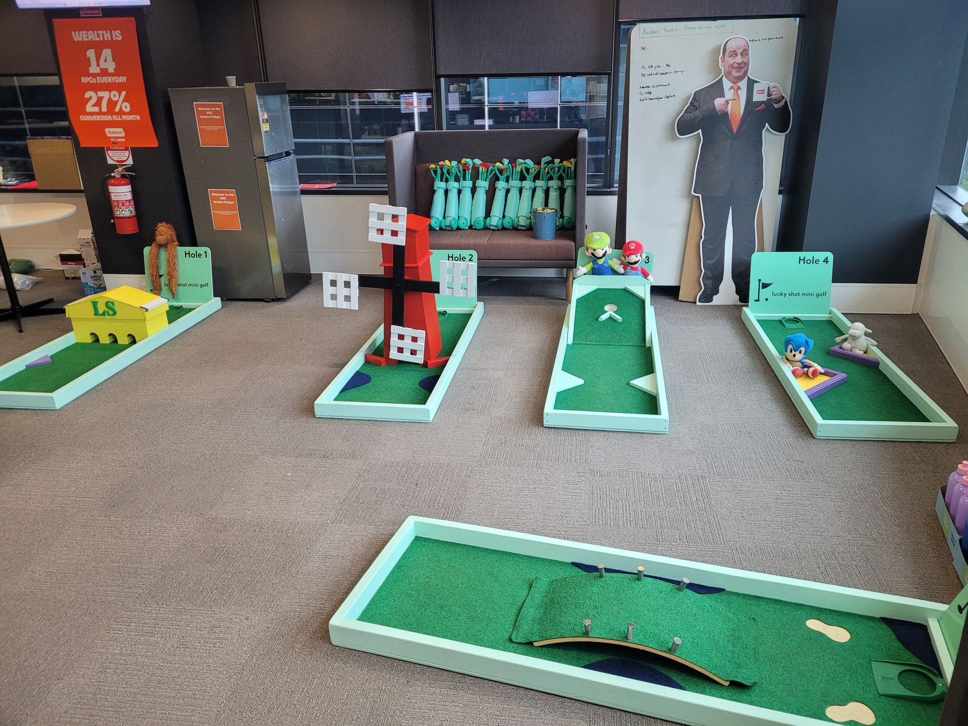 Indoor miniature golf course set up on carpeted floor, with windows overlooking a landscape.
