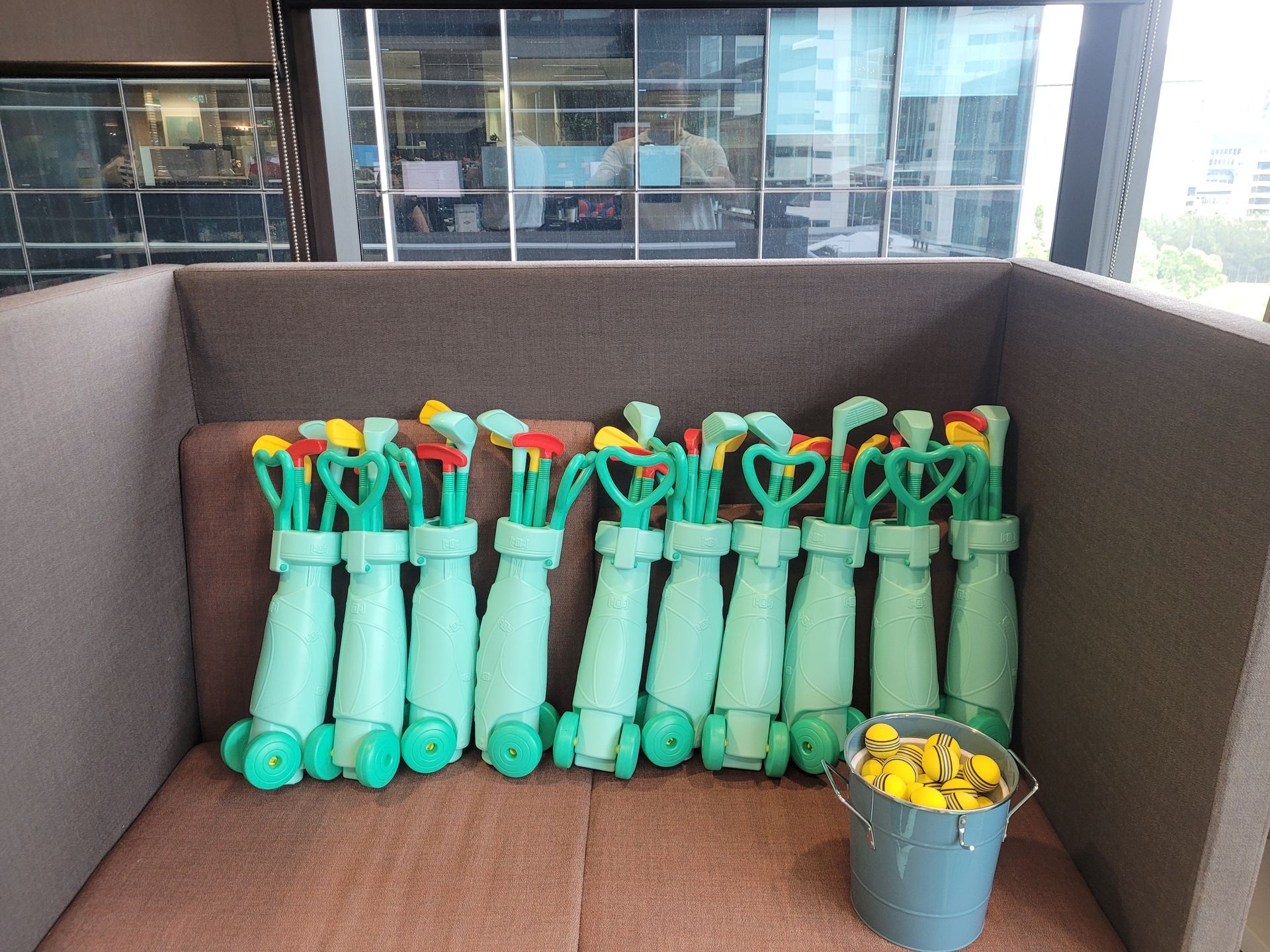 Row of teal toy golf club sets with yellow golf balls in a bucket, in a brown seating area.