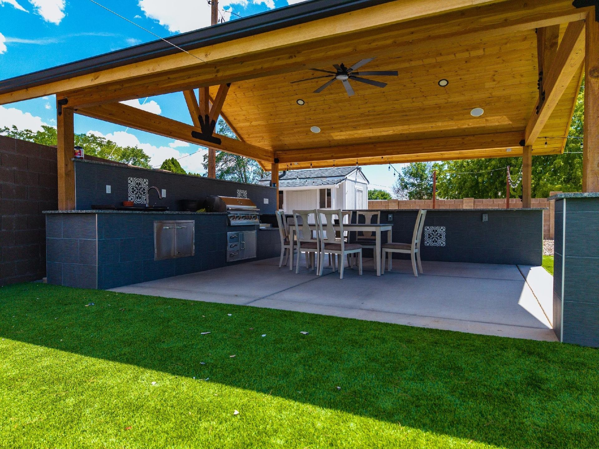 Outdoor kitchen and dining area under a wooden pergola, on a green lawn.