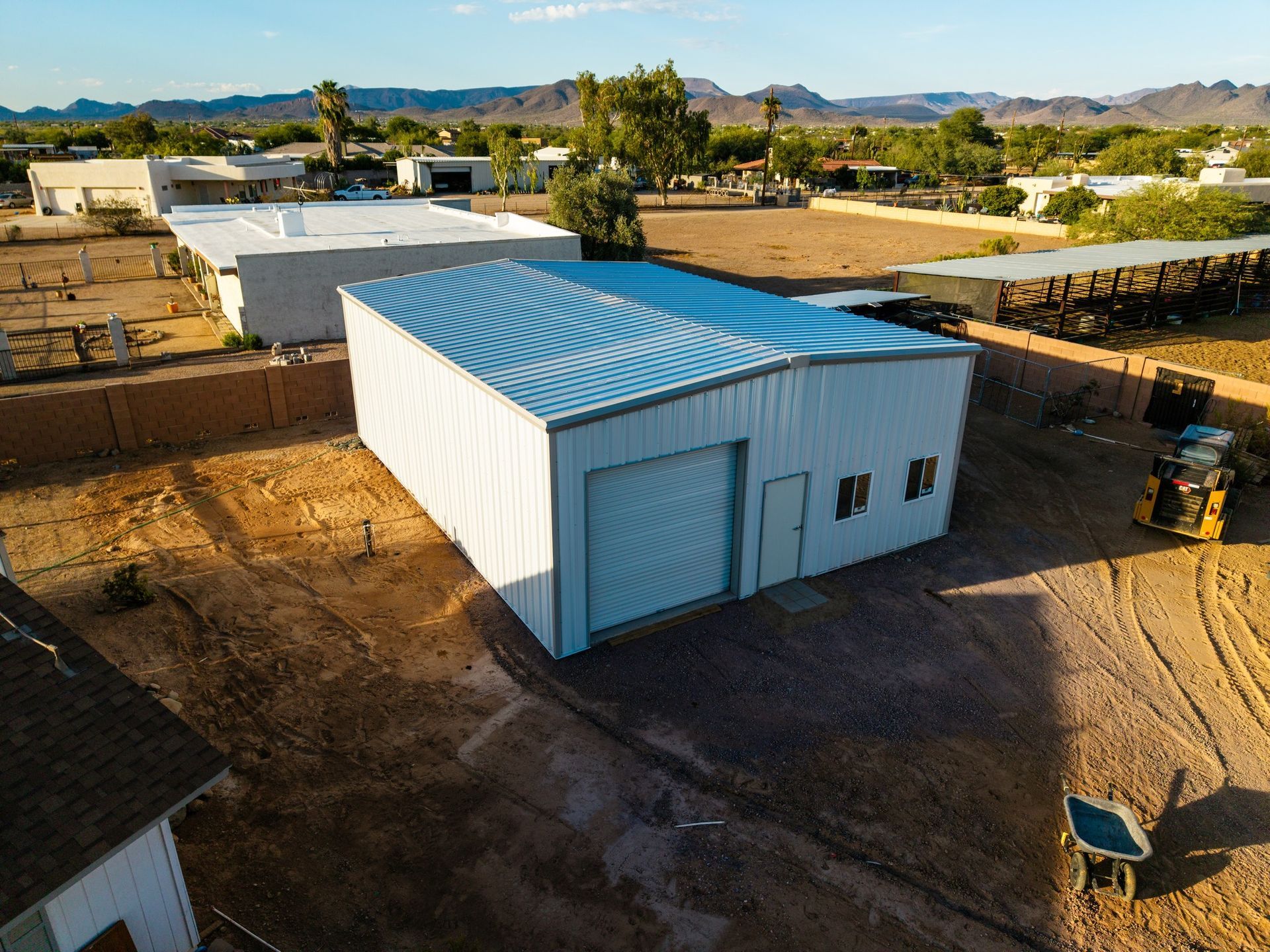 White metal shed with a corrugated roof in a desert-like setting, surrounded by dirt and other buildings.