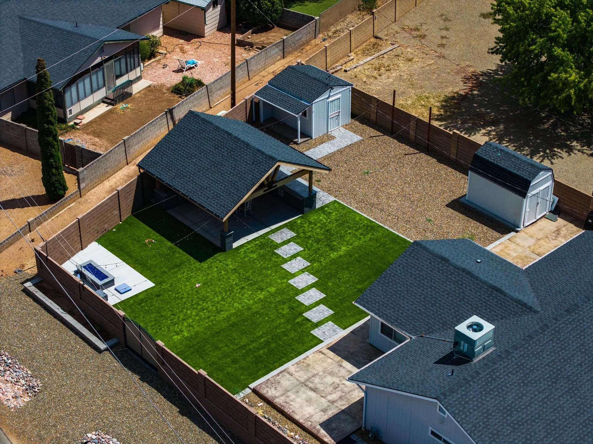 Aerial view of a backyard with green turf, a patio, sheds, and a house with a dark roof.