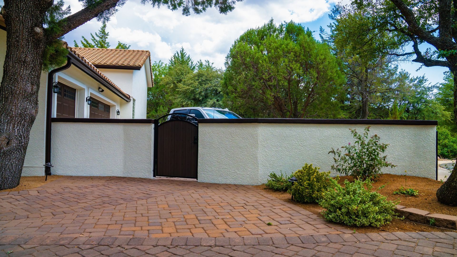 Brick driveway leads to white stucco wall with brown gate and vehicle visible behind it.