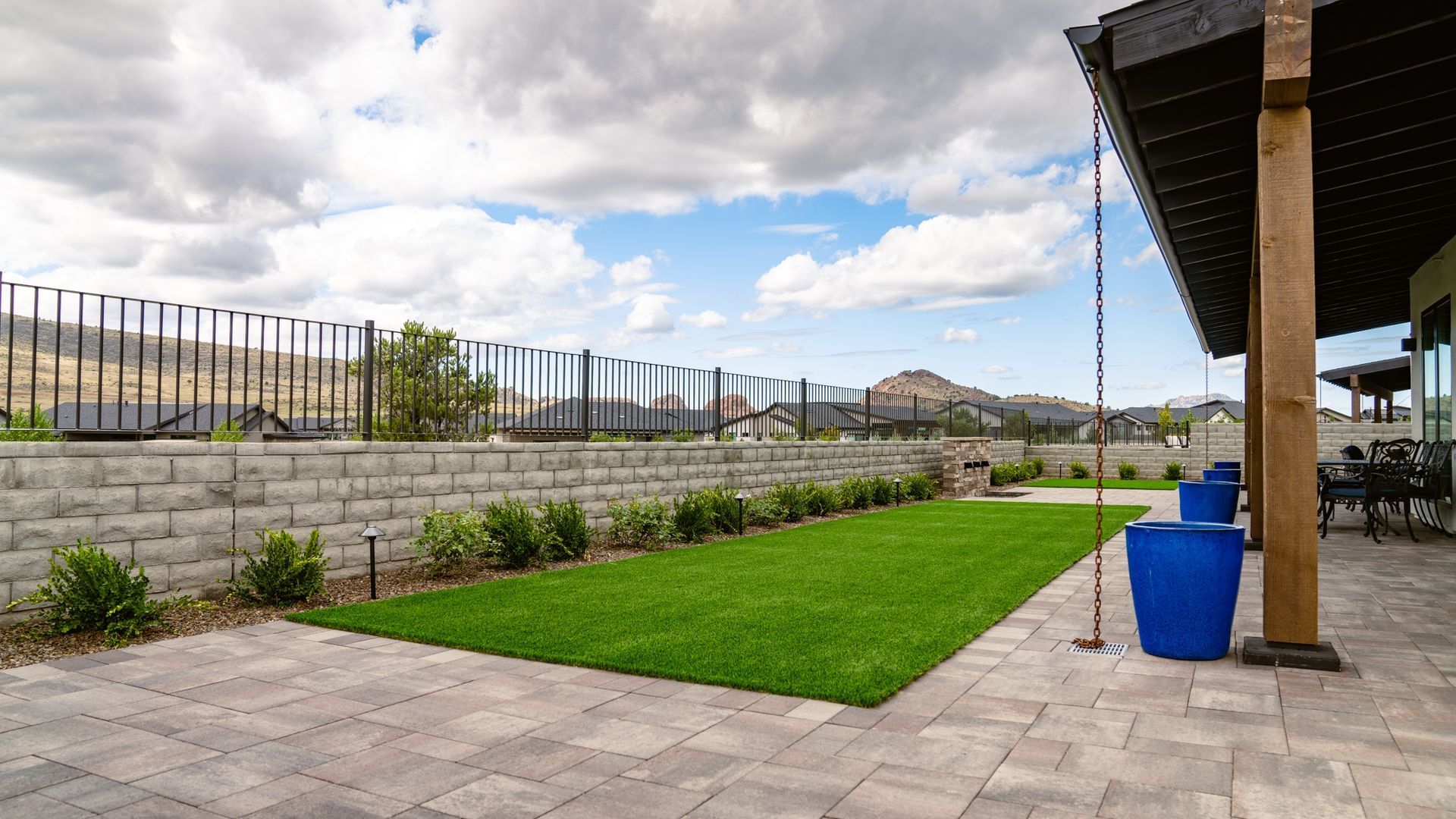Patio with brick pavers, green lawn, retaining wall, fence, and cloudy sky.