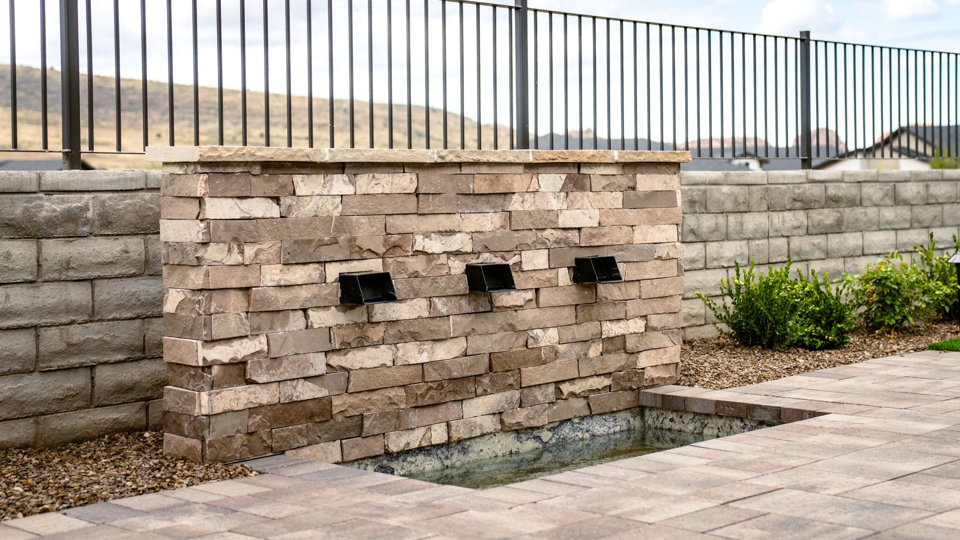 Brick wall water fountain with three spouts over a rectangular basin, set in a paved patio.