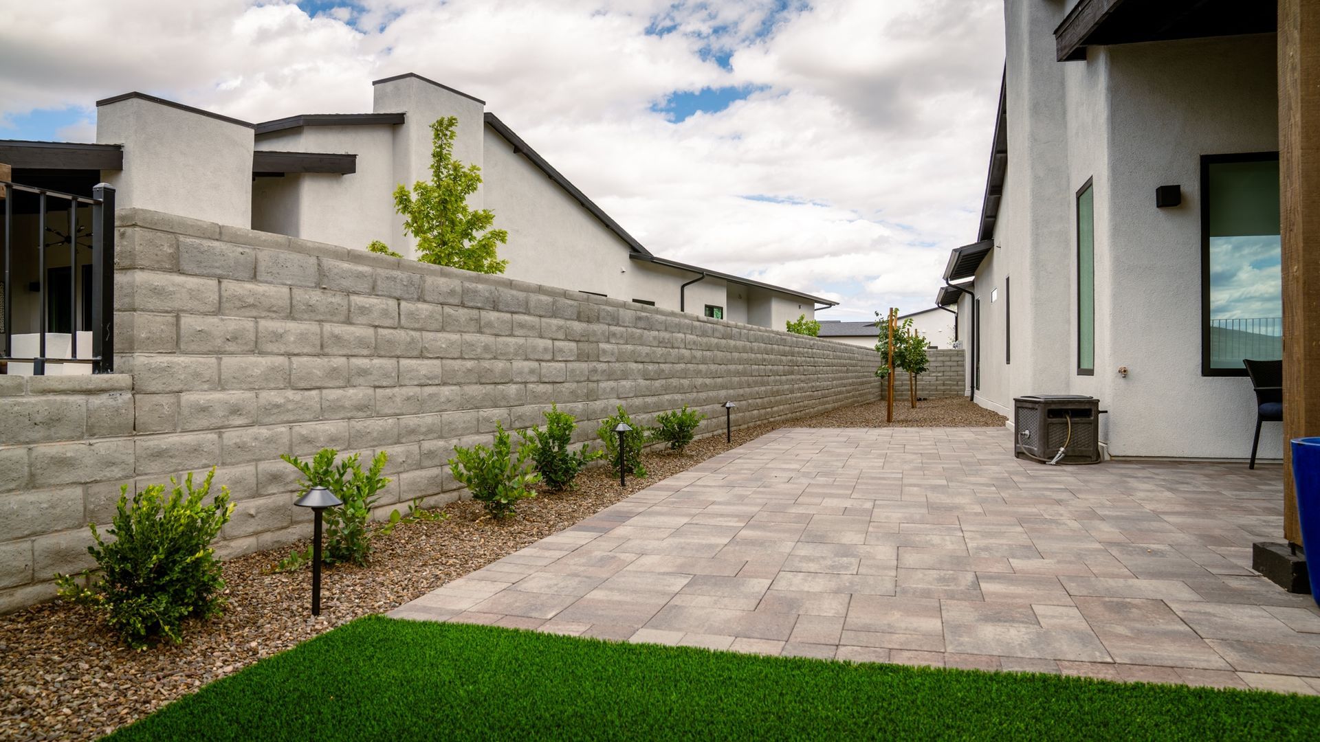 Patio with stone pavers, retaining wall, and green turf lawn. Bright white house and blue sky.