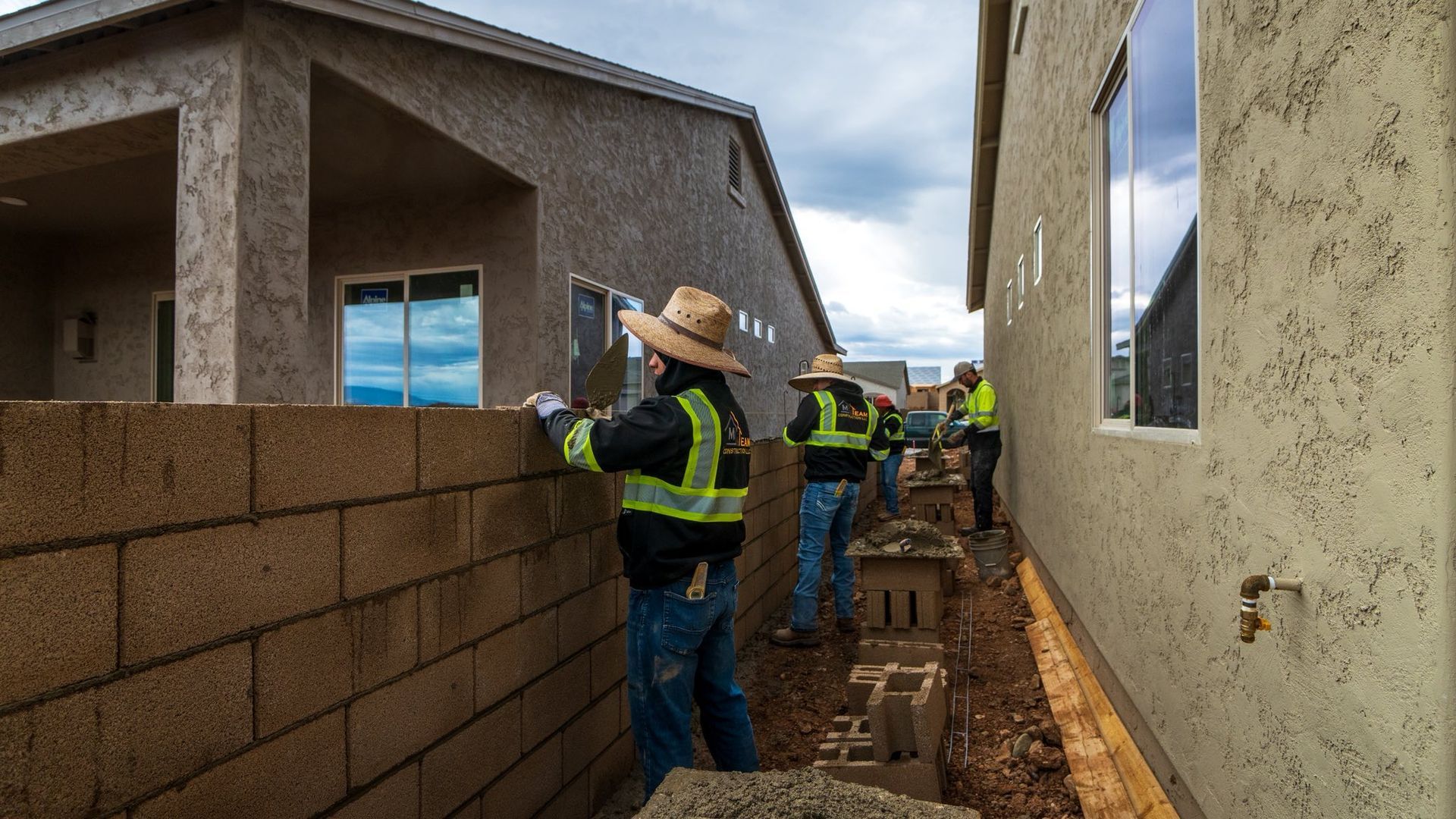 Construction workers building a brick wall between two houses; cloudy sky.
