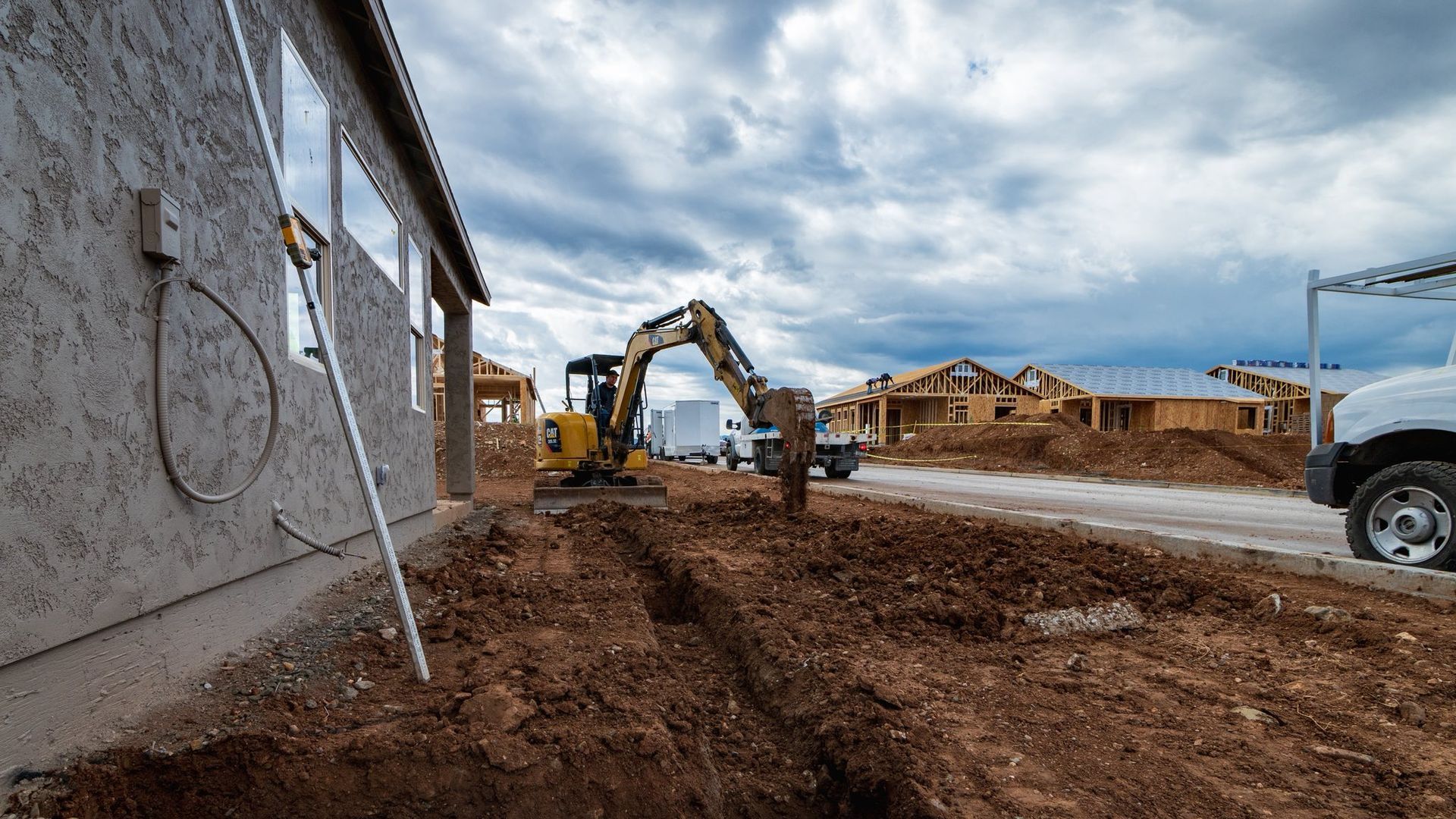 Construction site with excavator digging trench next to a stucco building. Houses under construction in the background under a cloudy sky.
