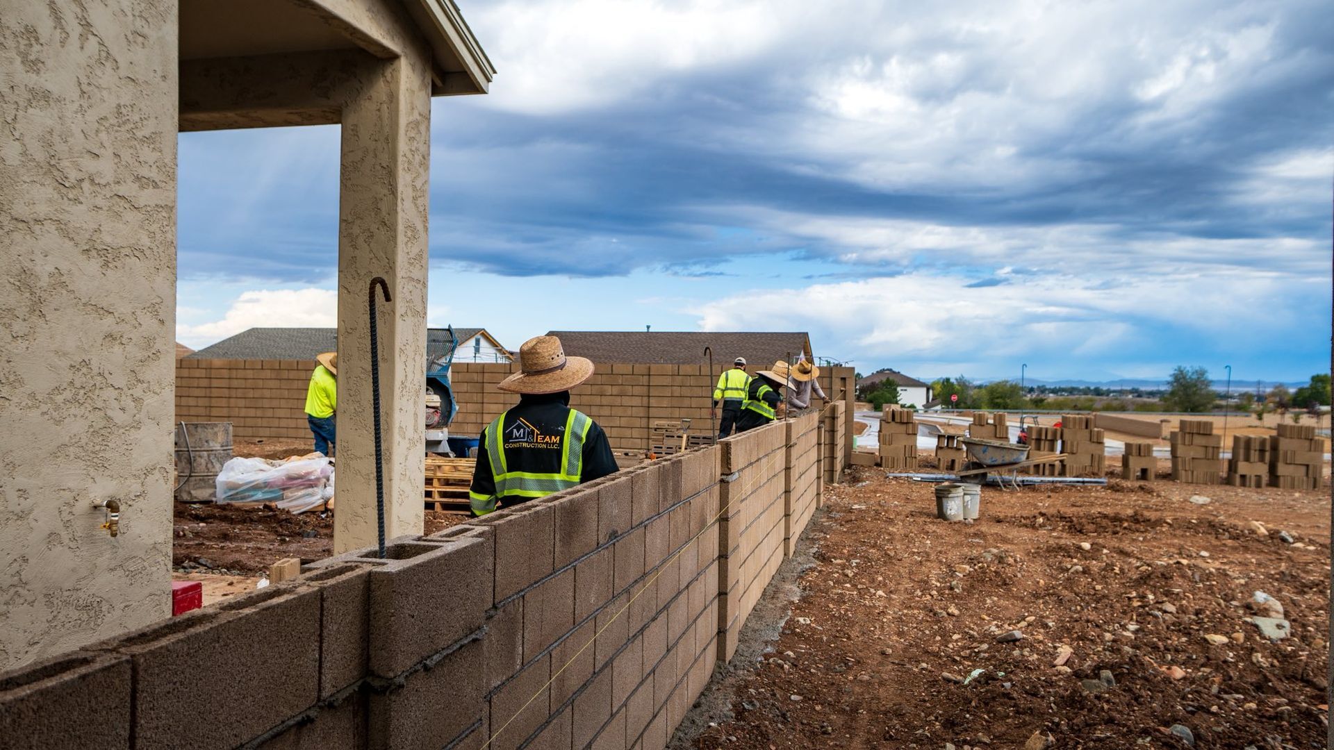 Construction workers building a cinder block wall, cloudy sky backdrop.