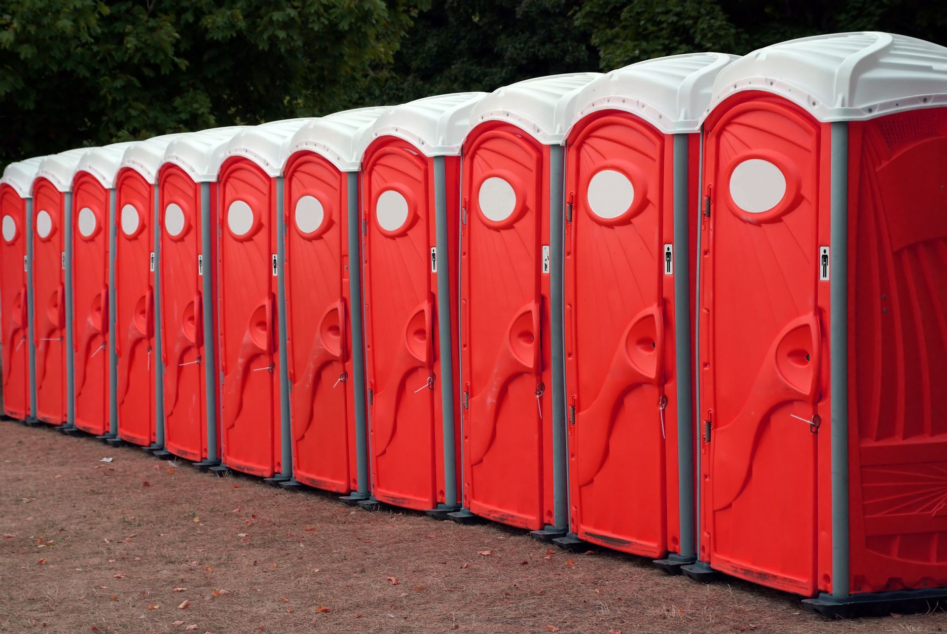 Long row of red portable toilets with white roofs lined up outdoors on dirt ground near trees.