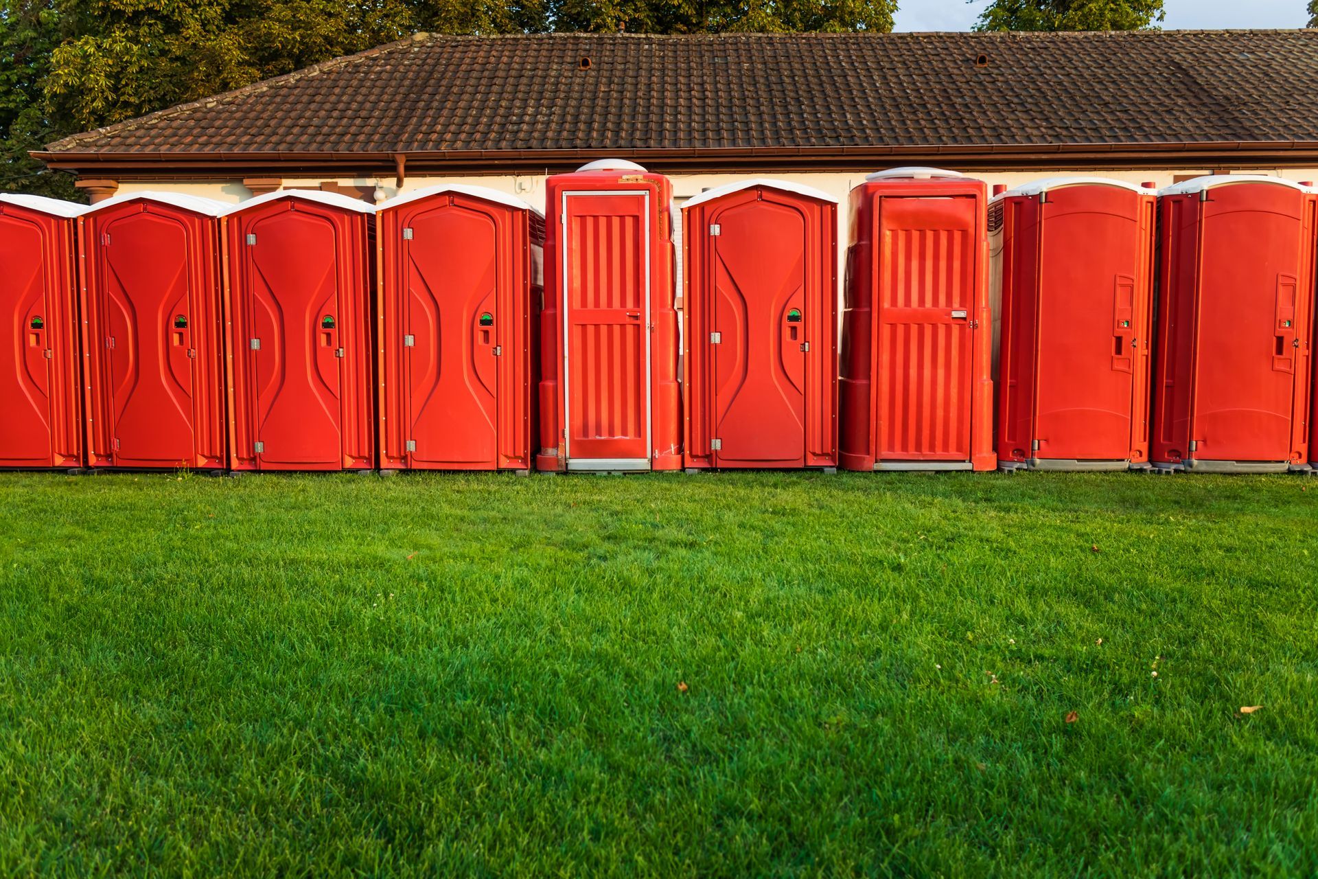 Row of bright red portable toilets lined up on grass in front of a building with a tiled roof.