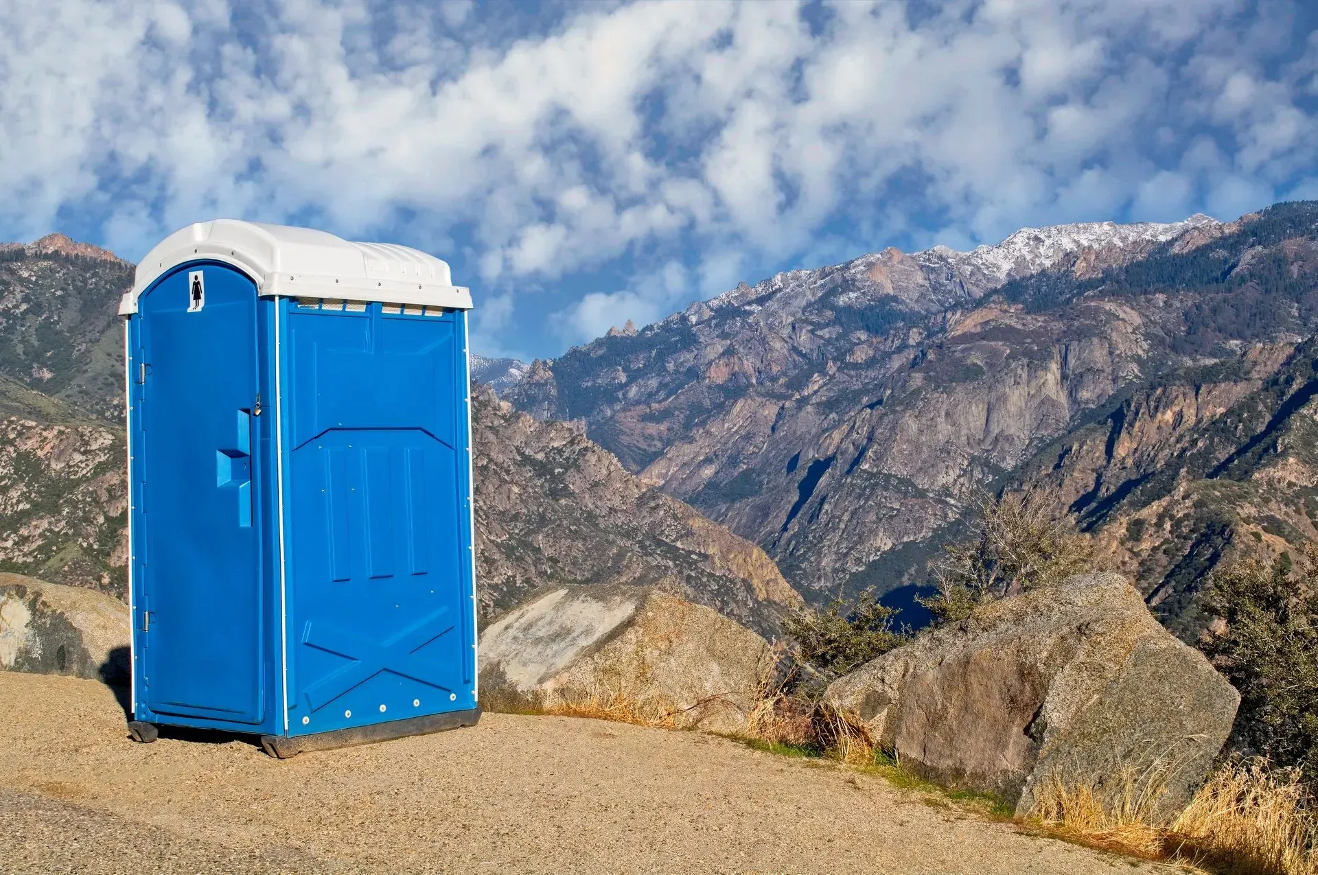 A blue portable toilet is sitting on top of a gravel road in front of mountains.