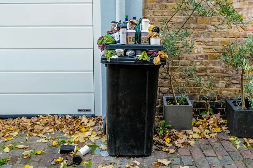 A black trash can filled with garbage is sitting on a brick sidewalk in front of a garage door