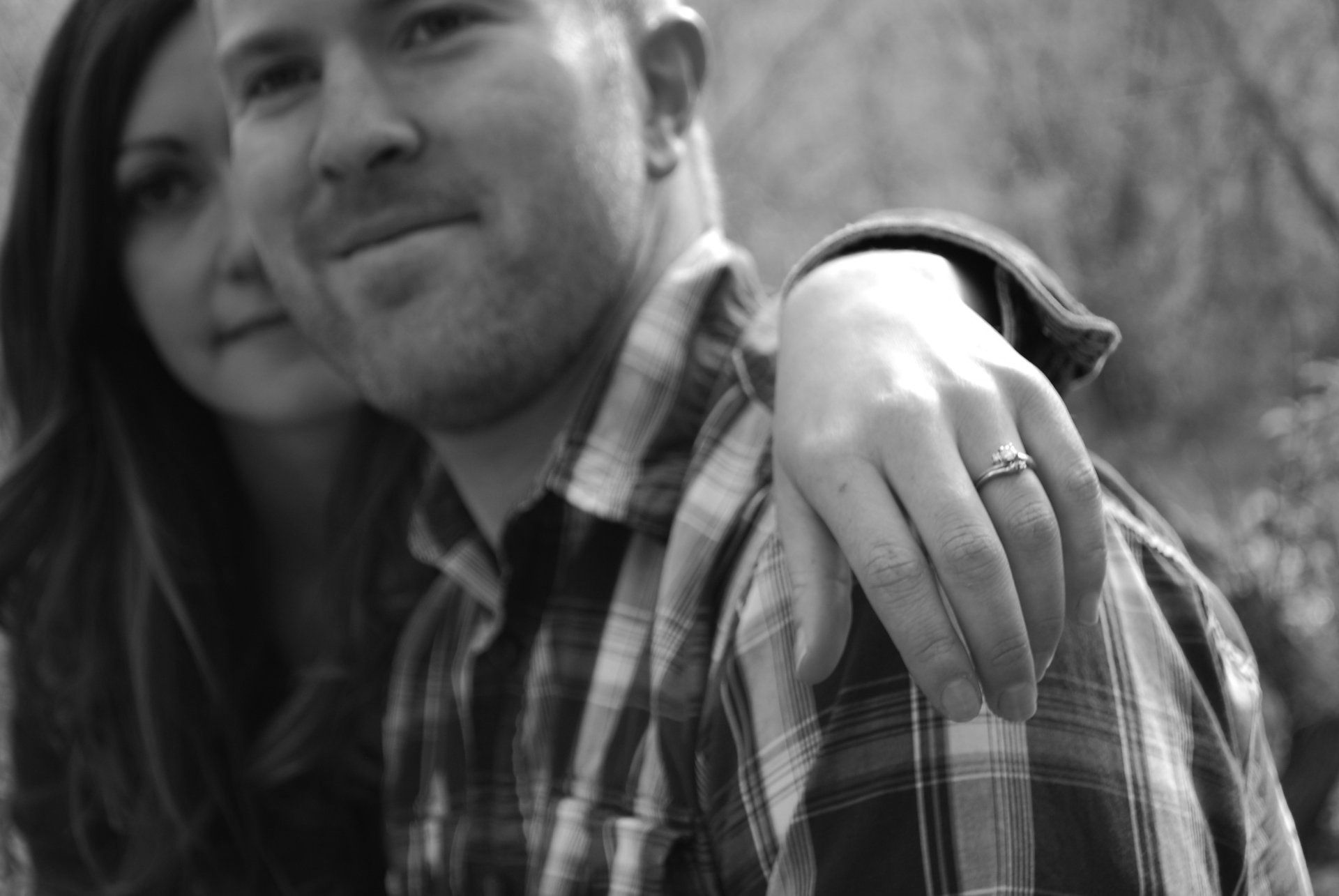 A man and a woman are posing for a black and white photo . the woman is wearing an engagement ring.