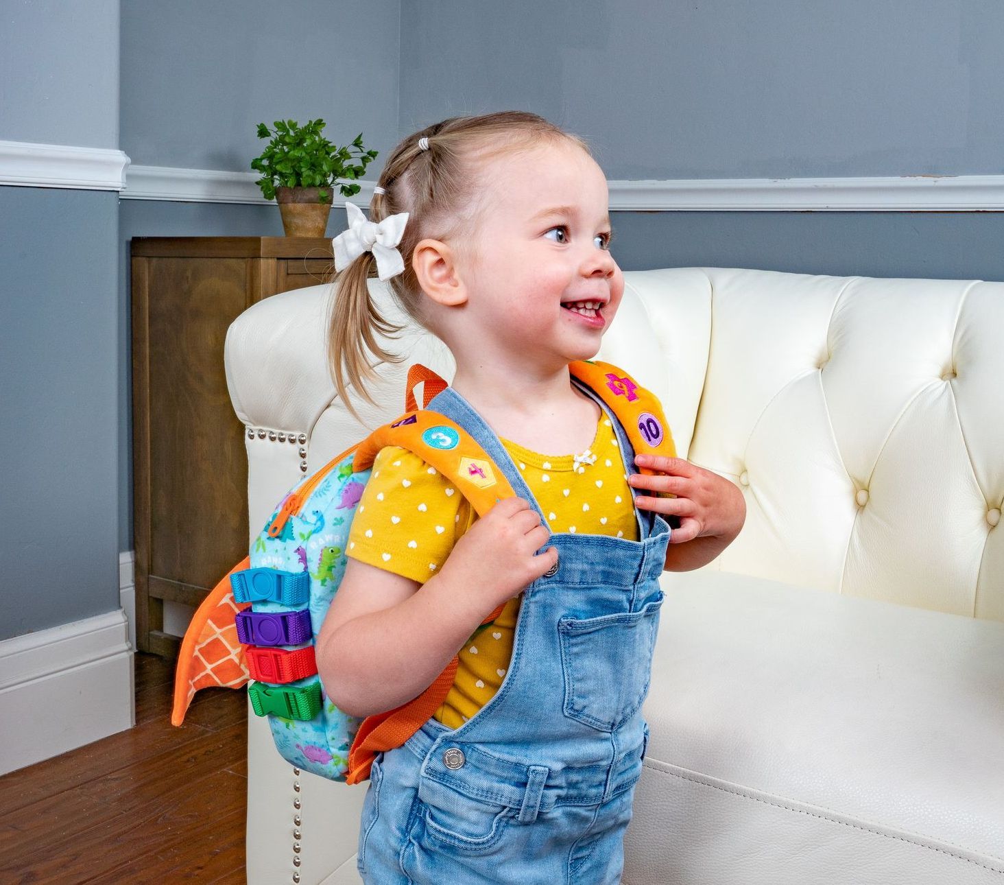 A little girl wearing overalls and a backpack is standing next to a couch.